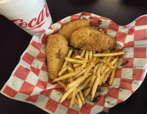 This image shows three chicken strips and French fries in a basket lined with paper. It also shows the bottom half or a cup for soda. Food from the grill in the Patio Café. Photo taken by The Signal reporter Marielle Gomez.