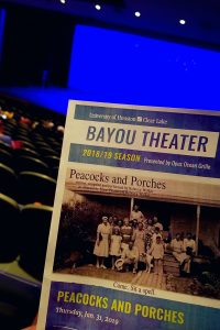 An image of the program for the Jan. 31, 2019 performance of "Peacocks and Porches" shown in front of the dimly lit Bayou Theater as audience members begin to take their seats.
