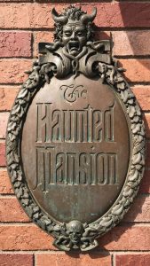 PHOTO: A zoomed in photo of the "The Haunted Mansion" sign. It is a copper sign that is rusting in areas,; a creepy face with snakes around it and a skull and snakes are also featured on the sign that hangs against a red brick wall. Photo by The Signal Managing Editor, Emily Wolfe.